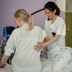 Ung pasient sitter i en seng med ryggen til - en står ved siden av sykepleier holder henne i hånden