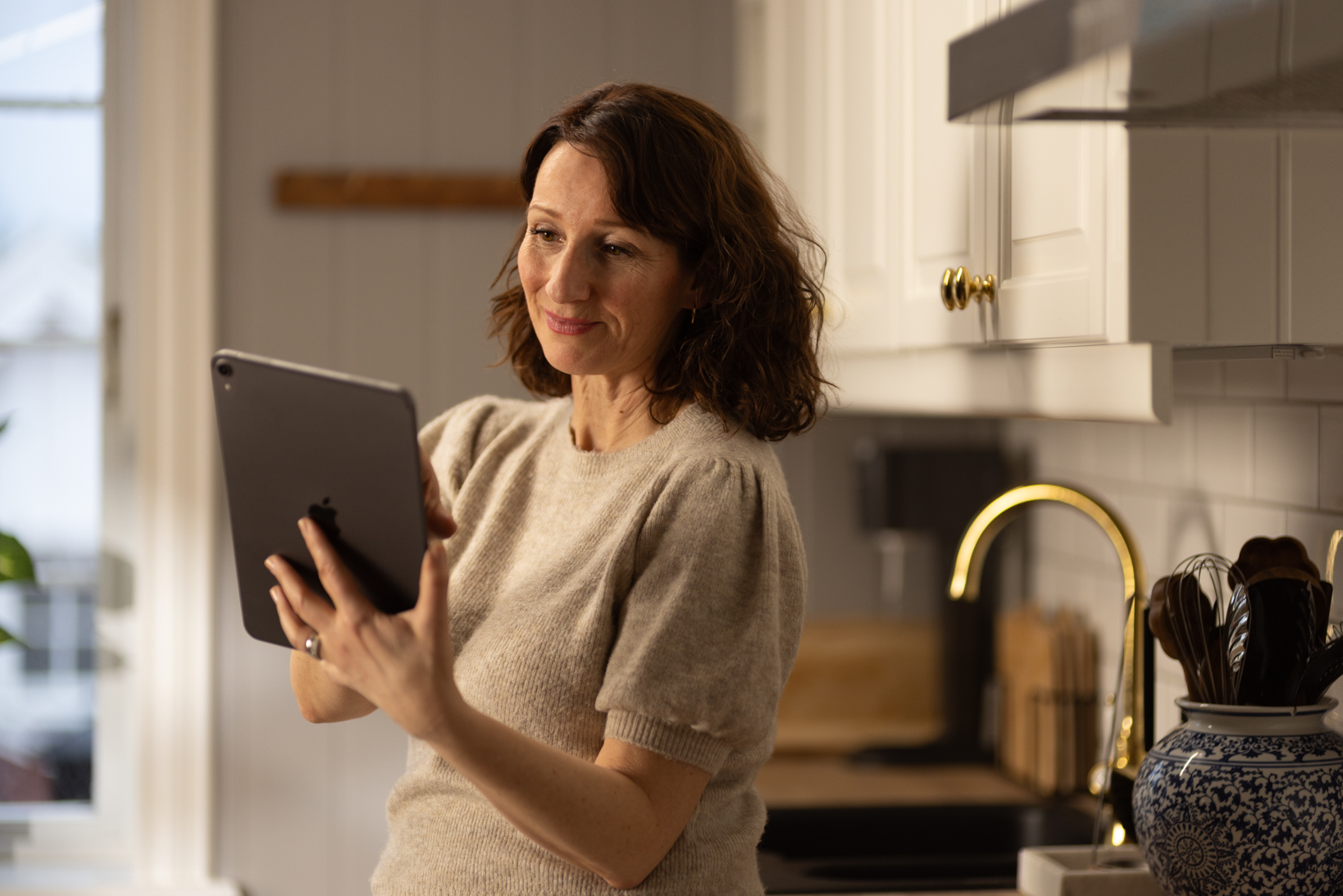 En kvinne står med nettbrett i hånden på et kjøkken.