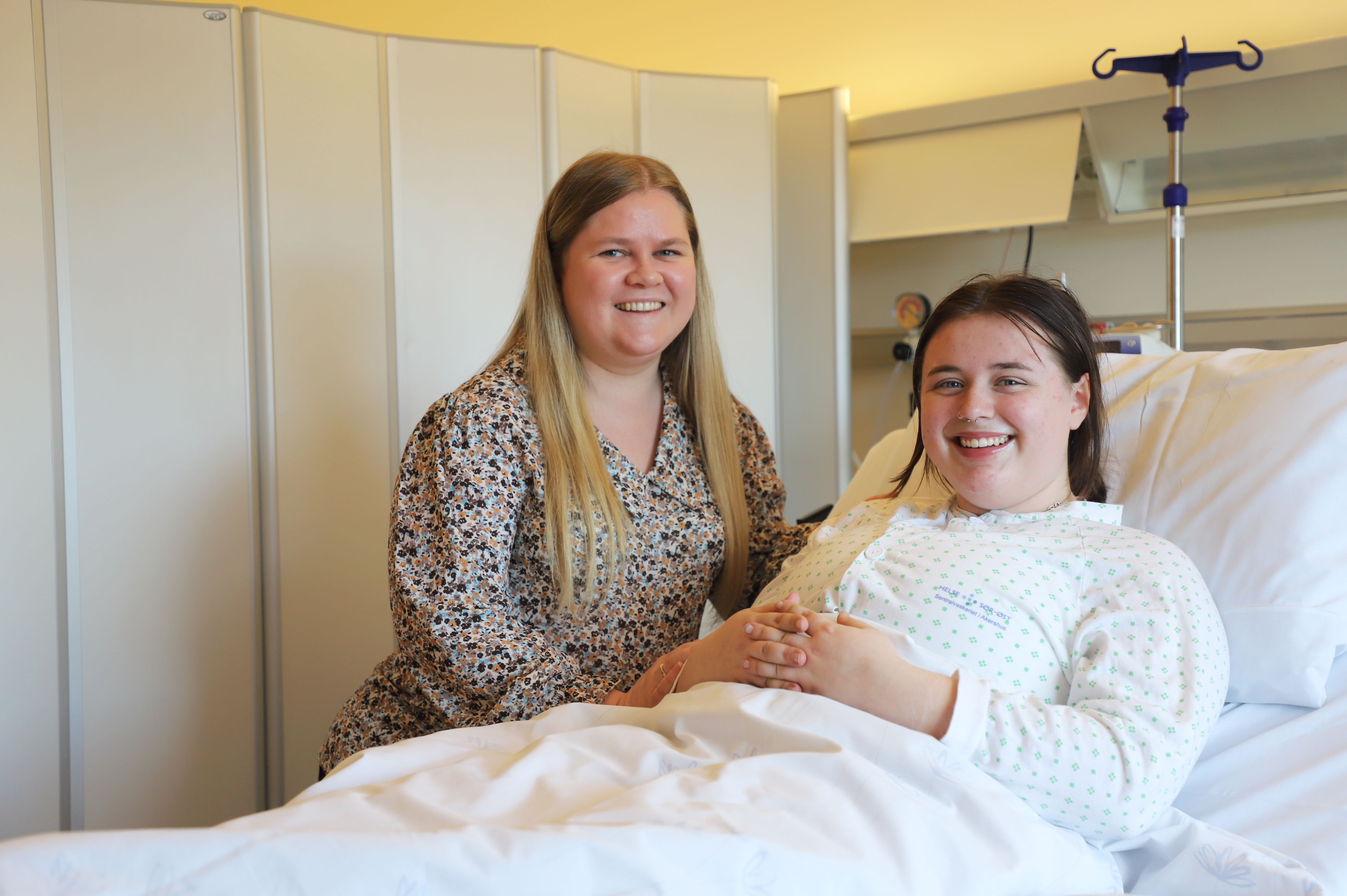 En jente ligger i en sykehusseng, og en venn står vedsiden av og smiler.