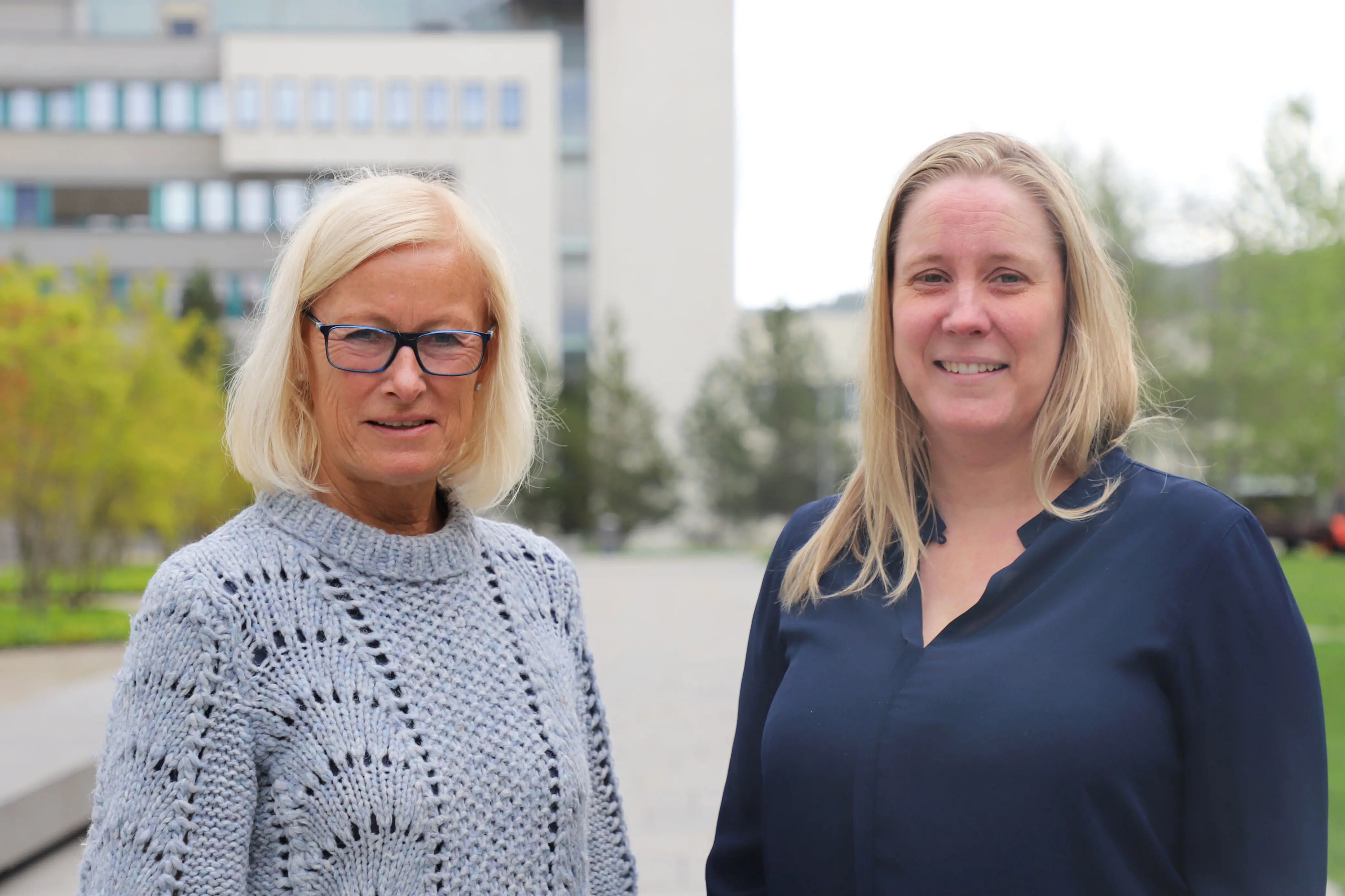 To kvinner foran Akershus universitetssykehus. Det er grønne trær i bakgrunnen. Kvinnene har på blå overdeler og smiler.
