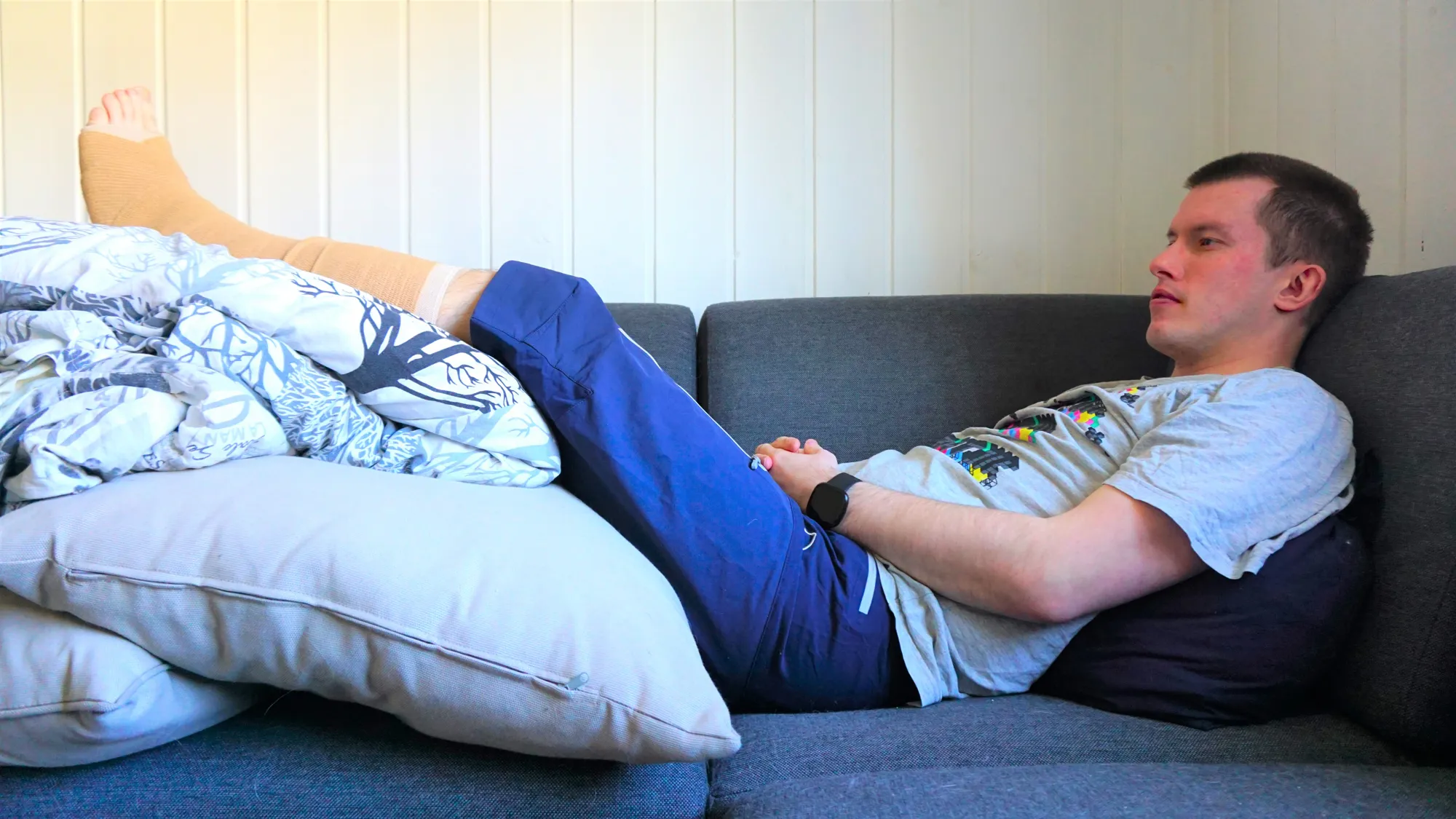 Mann sitter i sofa med gipset ben. Benet holdes høyt ved hjelp av puter. 
