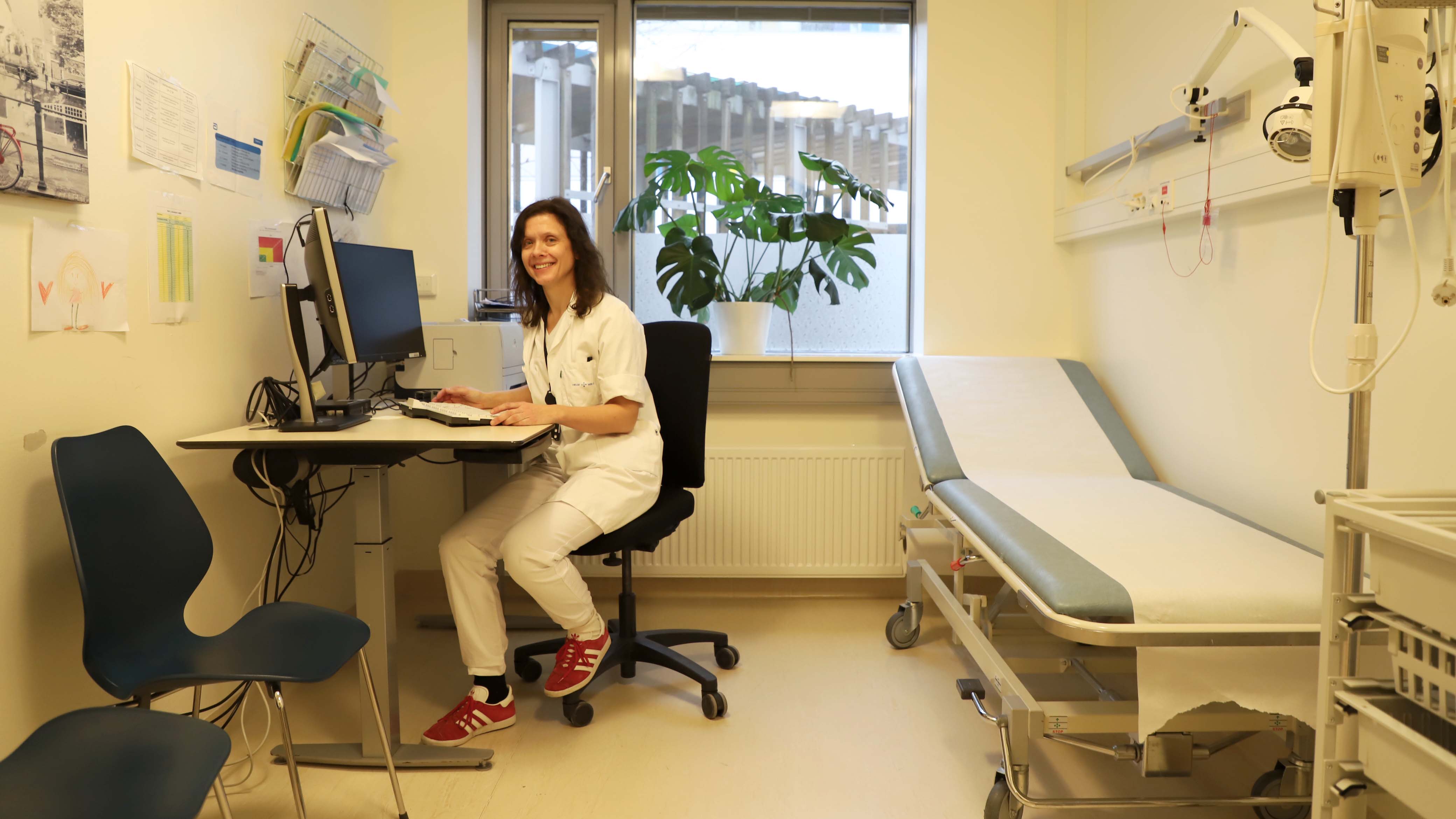 En blid kvinne sitter på kontorstol inne på et legekontor 