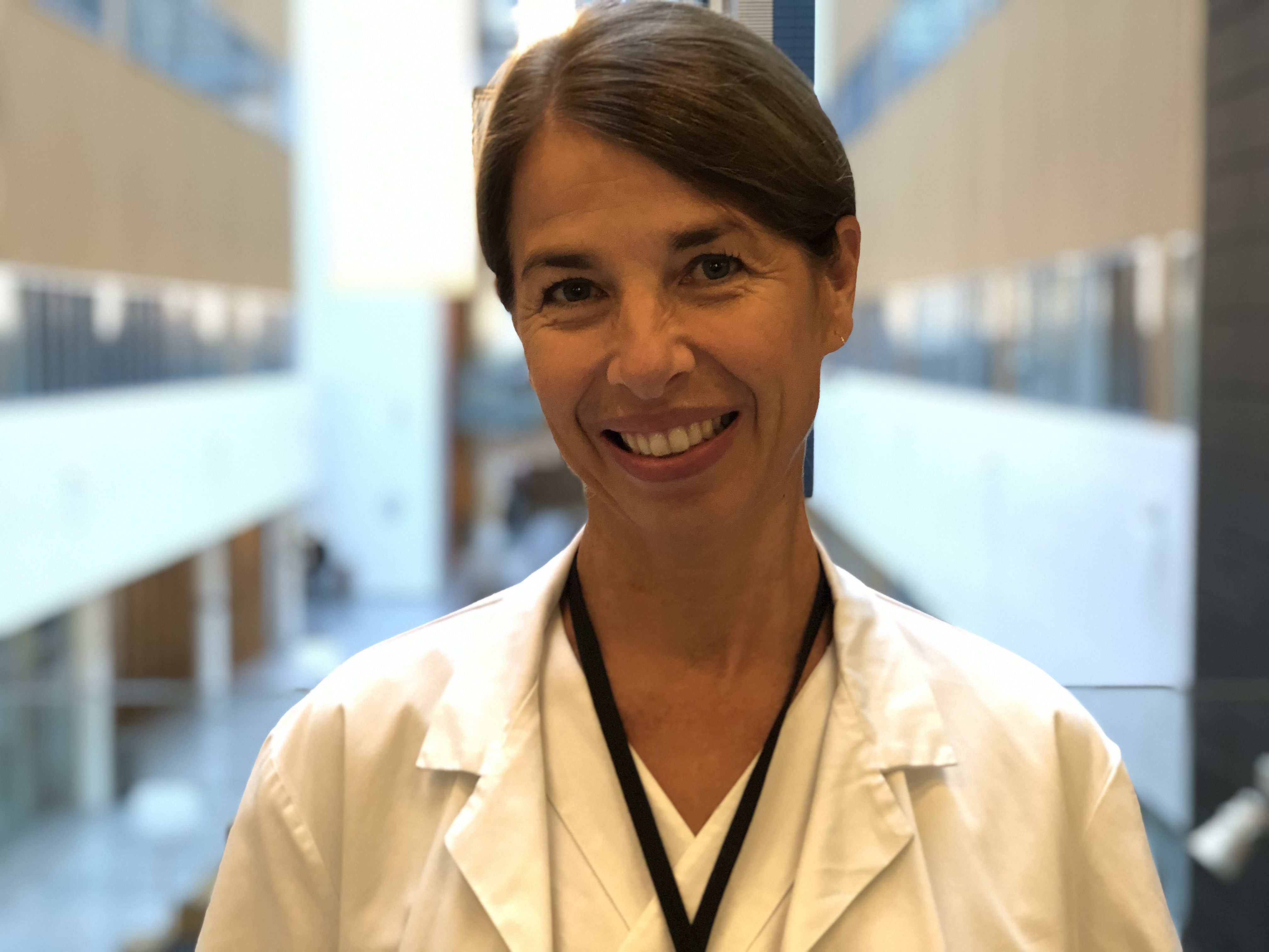 En kvinne med brunt hår i hestehale og hvit legefrakk smiler