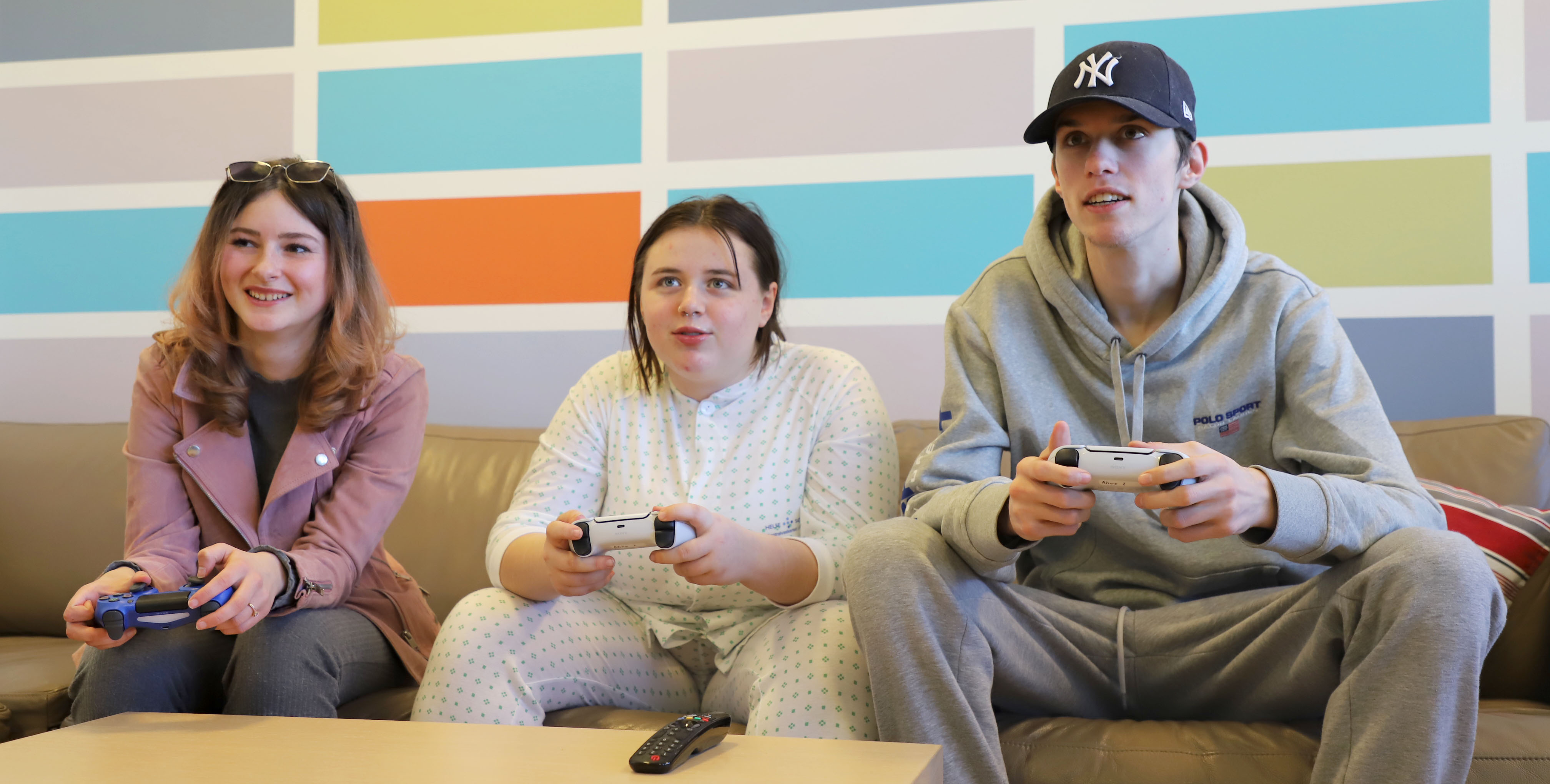 Tre ungdommer sitter i en brun sofa og spiller med hver sin playstationkontroll i hånden
