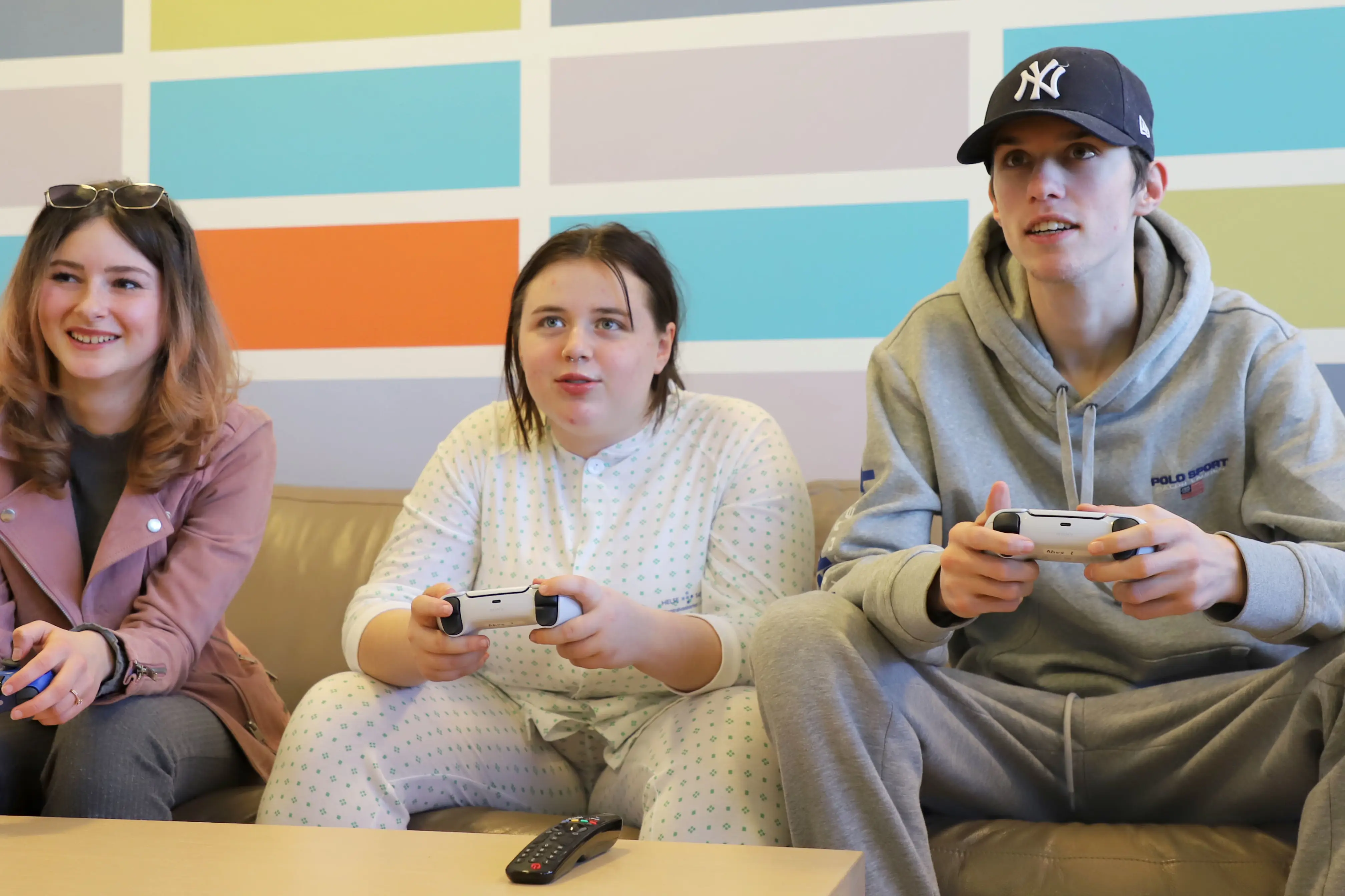 Tre ungdommer sitter i en brun sofa og spiller med hver sin playstationkontroll i hånden