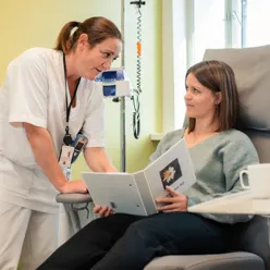 Pasient i stol får info fra sykepleier - pasienten holder i en brosjyre,