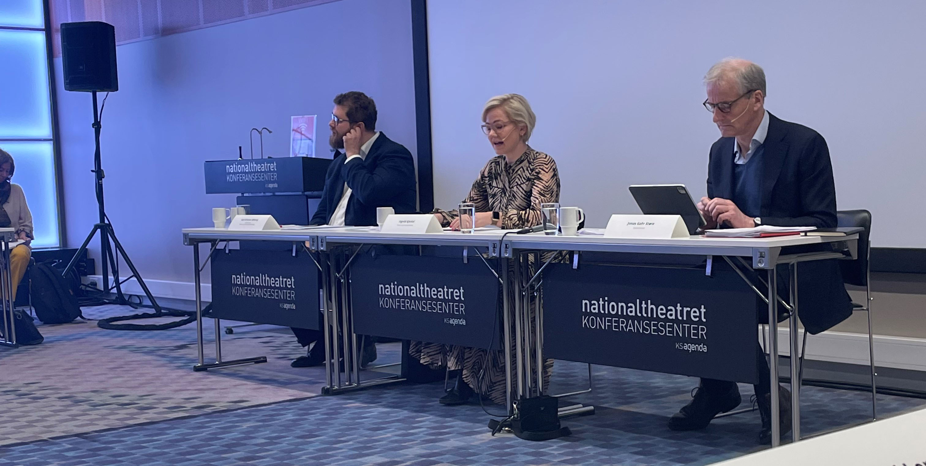 to menn og en kvinne sitter langs en bord. Ned fra bordene henger bannere hvor det står nationaltheatret konferansesenter.