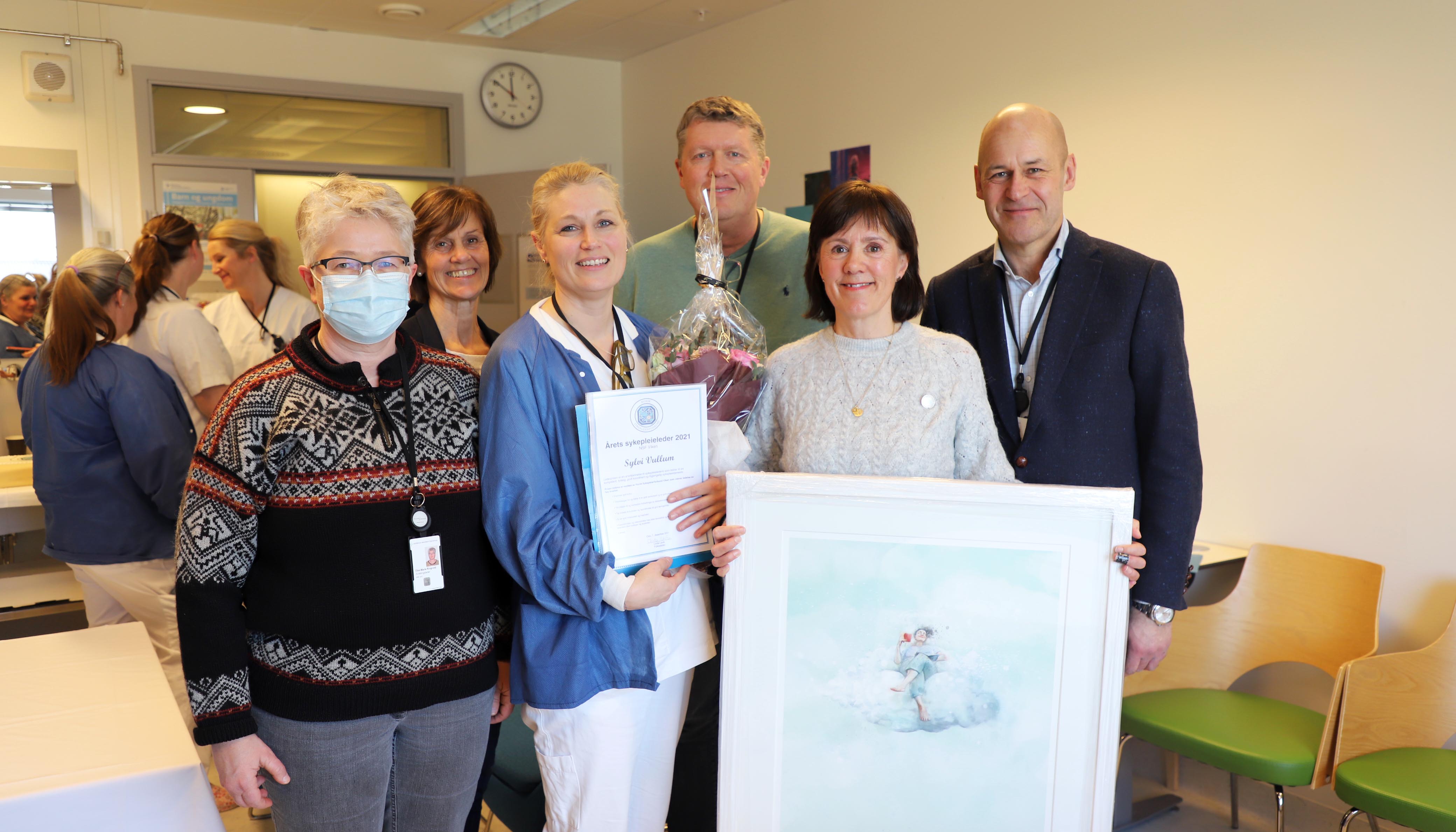 En gjeng med blide mennesker smiler til kamera. I midten står Sylvi Vullum med diplom for årets sykepleierleder.
