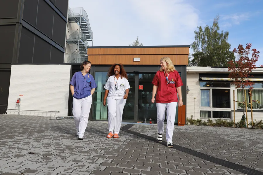 Fysioterapeut, sykepleier og ergoterapeut smiler foran sykehusbygg.