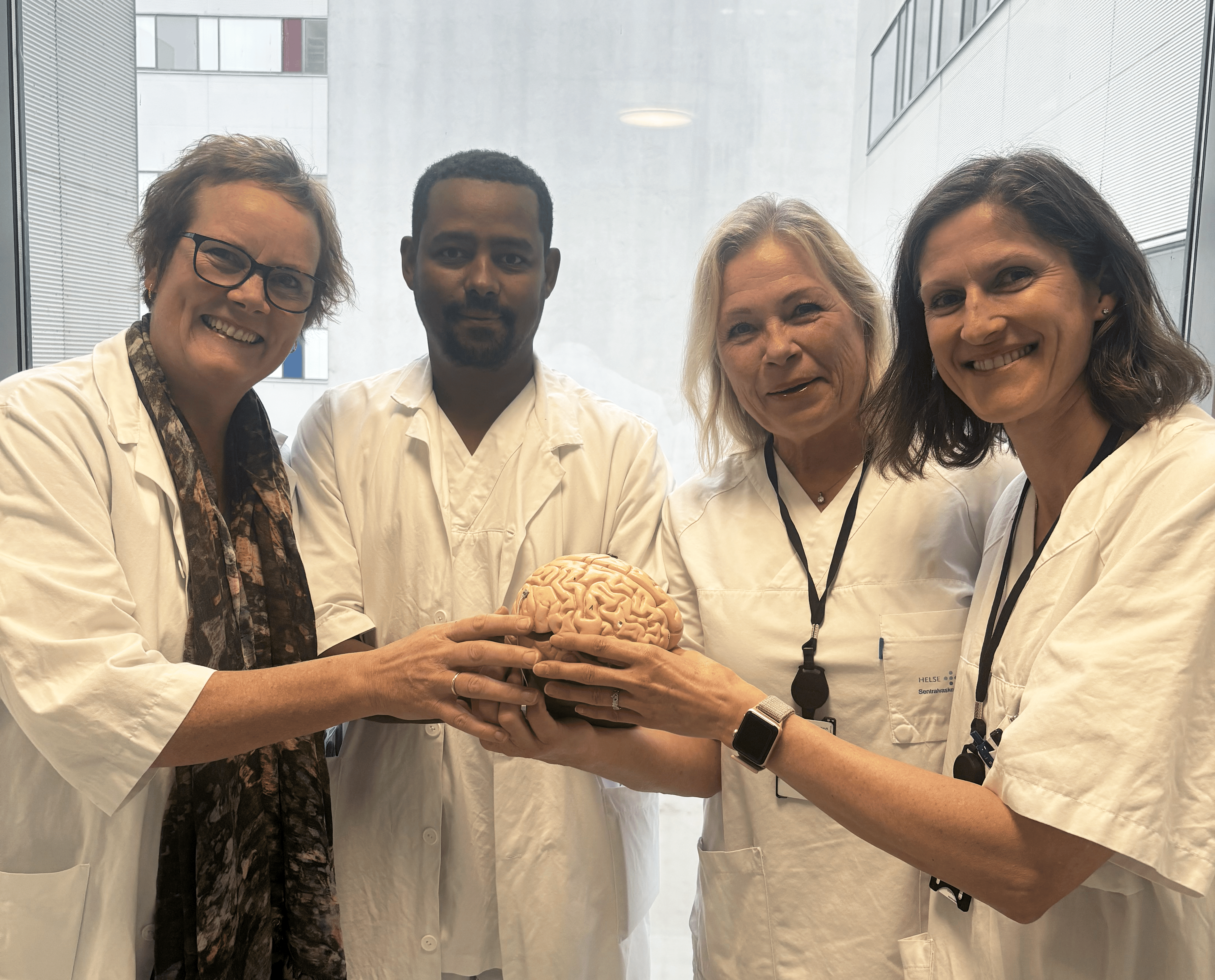 Fire sykehusansatte, to leger og to sykepleiere i sykehusklær holder en modell av en hjerne, ser i kamera og smiler
