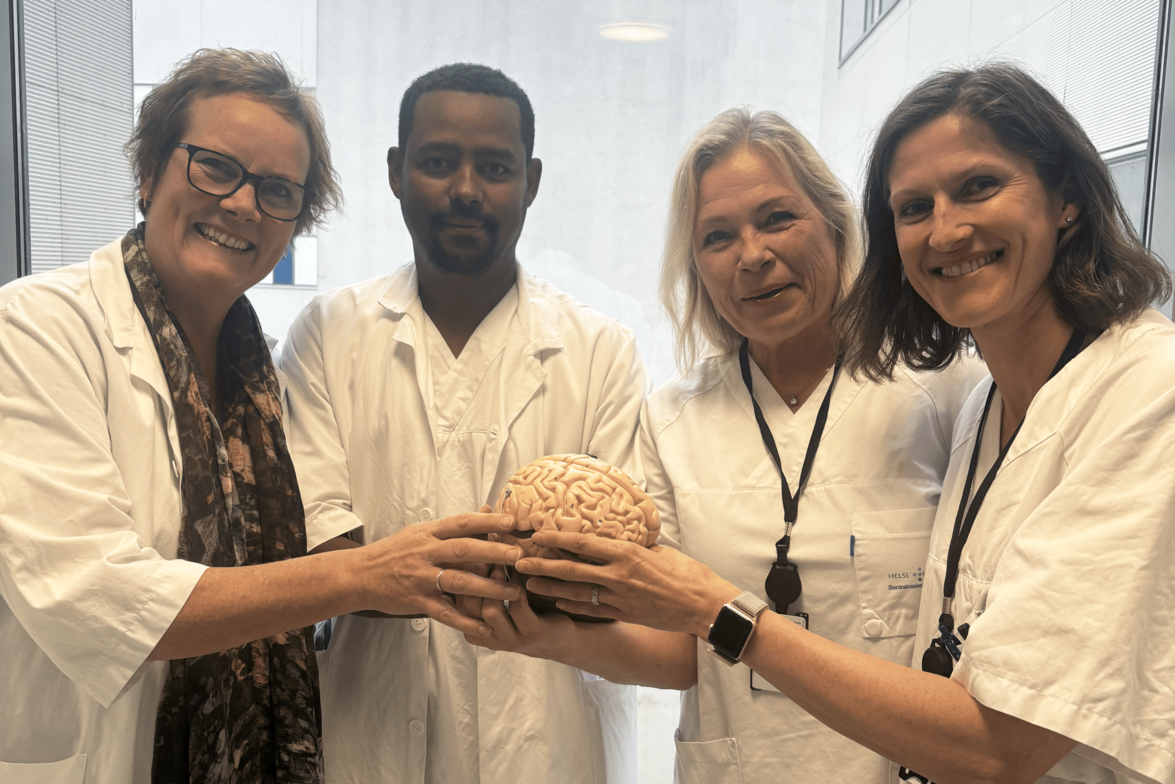 Fire sykehusansatte, to leger og to sykepleiere i sykehusklær holder en modell av en hjerne, ser i kamera og smiler